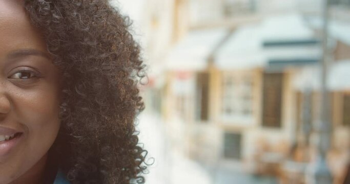Close up shot of African American young woman with curly hair looking at camera outdoors and smiling in good mood. Half face with smile on face. Happy female with positive emotions