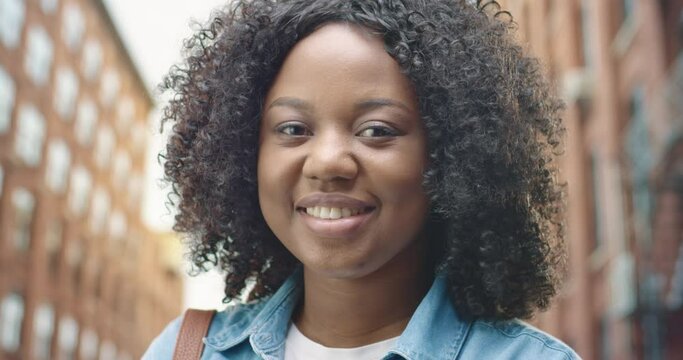 Close up of cheerful African American young pretty woman with curly hair standing on street, looking at camera and smiling in good mood. Beautiful female with smile on face. Positive emotions concept
