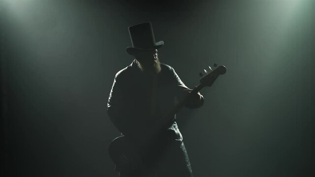 A bearded rock bass player plays a solo part during a studio performance with smoke and neon lights. Silhouette of a man in a hat and a long leather cloak plays the guitar. Close up.