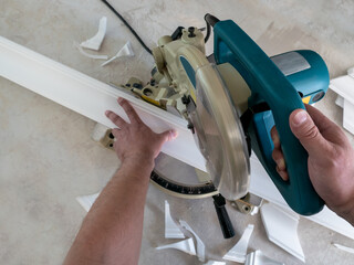 A worker cuts a ceiling molding made of polystyrene with a miter saw, close-up, first-person view. Cutting skirting boards with a professional power tool.