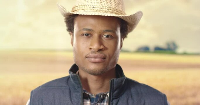 Close up portrait of African American young joyful guy in hat looking at camera in good mood standing in field outdoor. Handsome male agricultor in countryside, rural area. Cultivator concept