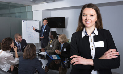 Obraz premium Portrait of confident positive young business woman in meeting room with working group behind