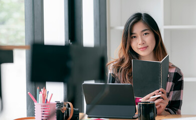 Portrait of young asian woman holding book and stylus pen, smiling and looking at camera while...