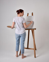 Full length portrait of a red haired artist girl wearing casual jeans and white shirt.  standing pose 
painting a canvas on an easel, against a studio background.