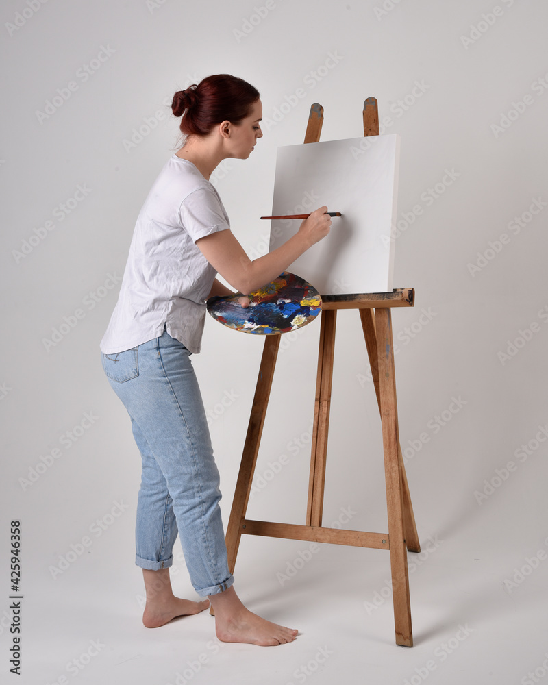 Canvas Prints Full length portrait of a red haired artist girl wearing casual jeans and white shirt.  standing pose 
painting a canvas on an easel, against a studio background.