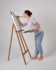 Full length portrait of a red haired artist girl wearing casual jeans and white shirt.  standing pose 
painting a canvas on an easel, against a studio background.