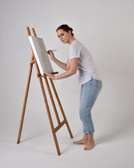 Full length portrait of a red haired artist girl wearing casual jeans and white shirt.  standing pose 
painting a canvas on an easel, against a studio background.