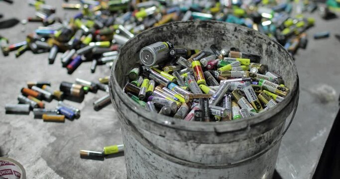 A full bucket of used batteries amid scattered piles of others. Processing and sorting. Environmental protection concept.