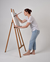 Full length portrait of a red haired artist girl wearing casual jeans and white shirt.  standing pose 
painting a canvas on an easel, against a studio background.