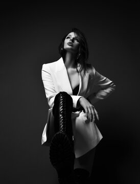 Black and white. Gorgeous naughty rich woman in jacket and high leather boot stands holding foot up, step on and looks up. Fashion, vogue, sexy stylish look for woman concept