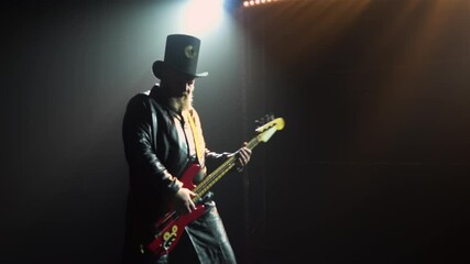 A bearded rock bass player plays a solo part during a studio performance with smoke and neon lights. Silhouette of a man in a hat and a long leather cloak plays the guitar. Close up.