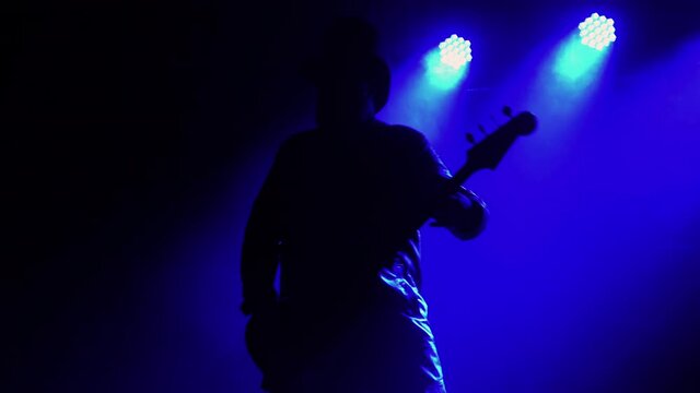 Active emotional musician playing bass guitar in dark smoky studio with blue lights. Silhouette of man in an original hat plays and moves to the beat of the music. Live rock concert. Close up.