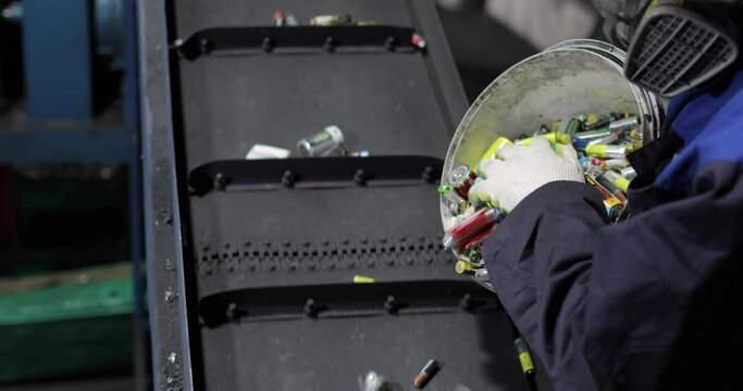 View of hands of person in uniform and gloves pouring out used batteries from bucket onto a moving conveyor belt, for separation and recycling. Processing. Environmental protection concept.