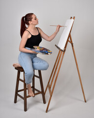 Full length portrait of a red haired artist girl wearing casual jeans and white shirt. Sitting pose on chair, painting a canvas on an easel against a studio background