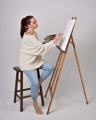 Fototapeta premium Full length portrait of a red haired artist girl wearing casual jeans and white shirt. Sitting pose on chair, painting a canvas on an easel against a studio background