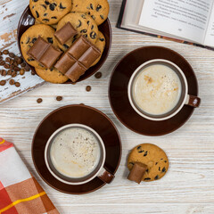 Two cups of coffee with chocolate chip cookies and chocolate on the background of an open book.