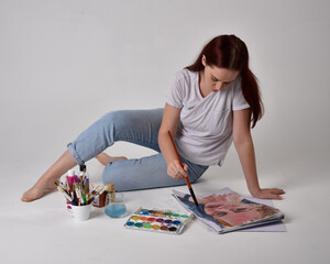 Close up  portrait of a red haired artist girl wearing white shirt.  Sitting on the floor 
painting with watercolour paints, against a studio background.