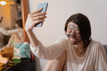 Woman doing black charcoal clay face cleansing mask at home in pajama, taking selfie with a phone, enjoying skincare rituals, having fun, Candid photo