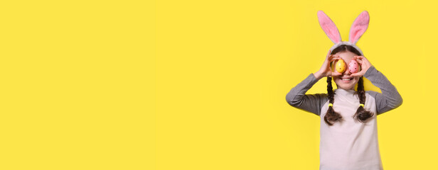 Funny happy girl with Easter bunny ears and colorful Easter eggs on a yellow background. Happy Easter! Copy space. Banner