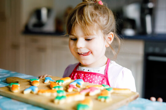 Cute little toddler girl and fresh baked homemade Easter or spring cookies at home indoors. Adorable blond child with apron with bunny and carrot cookie in domestic kitchen. Child eating cookie