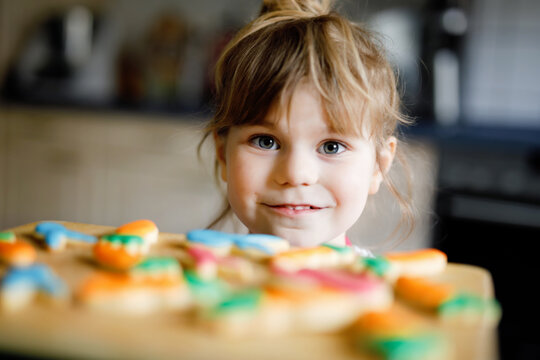 Cute Little Toddler Girl And Fresh Baked Homemade Easter Or Spring Cookies At Home Indoors. Adorable Blond Child With Apron With Bunny And Carrot Cookie In Domestic Kitchen. Child Eating Cookie