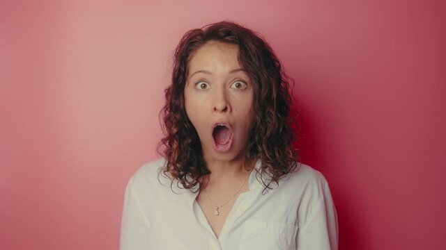 Shocked woman face with opened mouth and eyes looking to camera on a pink wall background. Portrait amazed girl with opened mouth in surprise