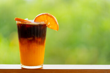 Americano coffee mixed with orange juice in a clear glass can be seen in layers. Behind it is a refreshing green bokeh.