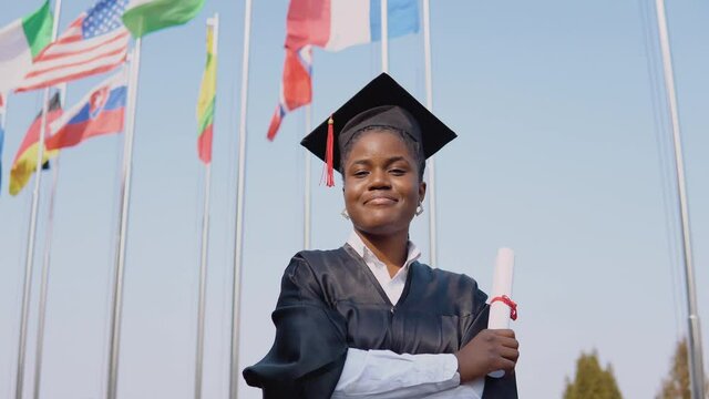 Young smiling african american female graduate standing in front of the camera with a diploma in her hands. The student stands outside with the international flags on background.