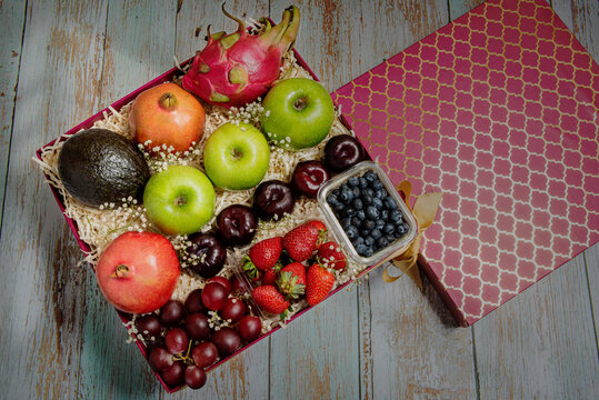 Fruit basket with avocado, plum, strawberry, blueberry, grapes, green apple, apple, guava, pomegranate, in a red box with a golden ribbon bow placed on a rustic table with window light from the side.