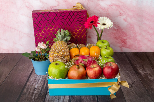 Fruit basket with gerbera flowers and pineapples, kiwi, orange, green apples, apple, dragon fruit, guava, pomegranate, in a blue and red box with a golden ribbon bow.