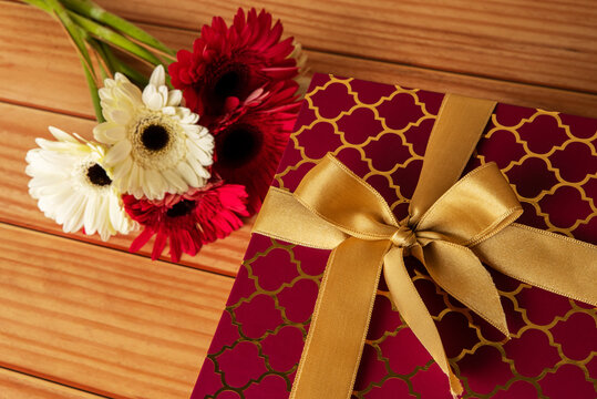 Red gift box with a golden ribbon bow on a wooden desk with flowers next to it