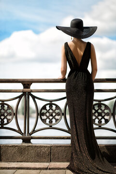 Woman Back Side View in Hat and Evening Dress Outdoor. Fashion Model Rear View looking away. Luxury Lady in long Gown at Promenade looking at Sky