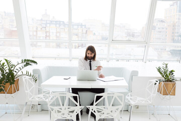 business man in a white shirt and tie sits at the table and works with documents and laptop in a modern office by the window
