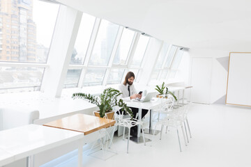 a business man in a white shirt and tie uses the phone and sits at a table with a laptop on the background of a white modern office by the window
