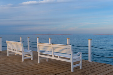 Wooden white benches at the pier with the view of blue sea, cloud beautiful sky, no people, lonely and calm feeling, picturesque view, copy space.