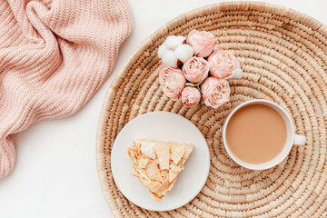 Wicker tray, cup of coffee with milk, piece of cake, rose flowers, pink knitted plaid or blanket. Breakfast in bed. Stylish home interior decor. Flat lay, top view.