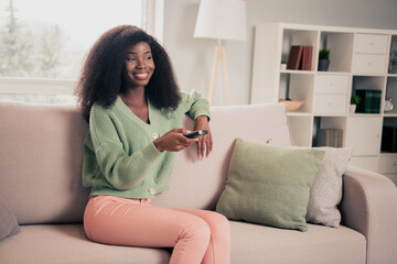 Photo of young attractive african woman happy positive smile sit couch home watch tv switch channel remote controller