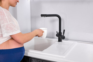 cropped pregnant woman stands near tap fills up water to drink after training, side view