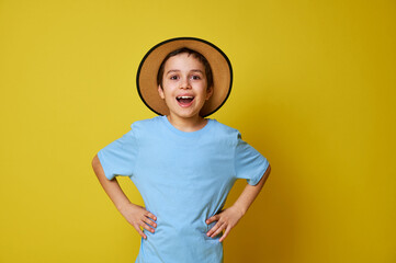 Happy boy wearing a summer hat and blue t-shirt, putting his hands on his waist and looking happily into the camera. Facial expressions, emotions on a yellow background with space for text