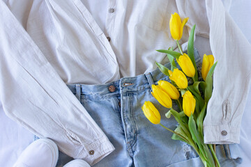 Women clothing flat lay with blue jeans shorts, beige shirt, white t shirt and yellow tulips, fashion blogger content, denim and linen texture