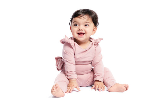 Cute female baby in lovely pink outfit smiling with mouth open and looking aside. Attractive little baby sitting on floor and posing, isolated on white studio background. Concept of childhood.