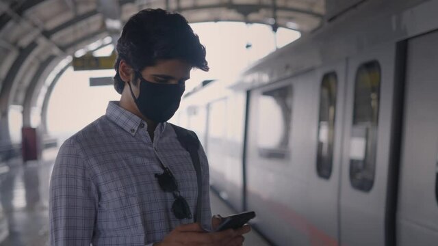 A Young Attractive Corporate Office Male Employee Is Standing At The Metro Or Subway Station And Using Mobile Phone To Type A Text Message Or Drafting A Mail While Train Passing In The Back 