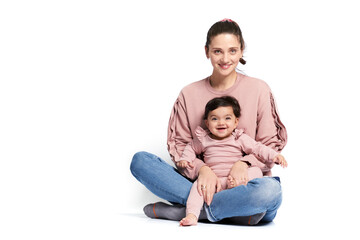 Fototapeta premium Portrait of cute mother with daughter looking at camera, isolated on white studio background. Young attractive woman holding sweet adorable child on leg while sitting on floor in lotus pose.