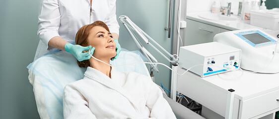 Adult woman getting rejuvenation skin face with ozone therapy procedure at beauty clinic using oxygen skin care machine - obrazy, fototapety, plakaty