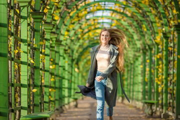 Fototapeta premium Beautiful happy young woman in a gray coat walks along a picturesque alley in an autumn park. Attractive woman with light brown hair smiles happily. Fall season. Amazing autumn mood.