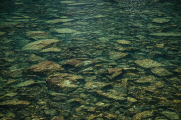 Meditative ripple and many reflected lights in green mountain lake. Beautiful relaxing background of stony bottom in turquoise transparent water of glacial lake in sunlight. Many stones under water.