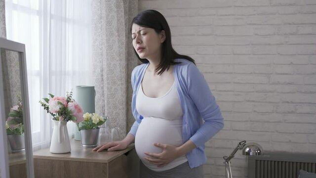 chinese pregnant mom approaching to window is touching her belly while suffering unexpected contraction pain in the bedroom.