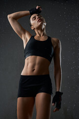 Fototapeta na wymiar Portrait of wet fit caucasian female model, wearing black sportswear and gloves standing, loft interior. Young muscular fitnesswoman wiping sweat off forehead after hard training under rain.