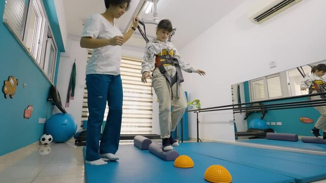 Wide shot of a physiotherapist helping a young girl do rehabilitation balance exercises while supported by a harness at a physiotherapy session.