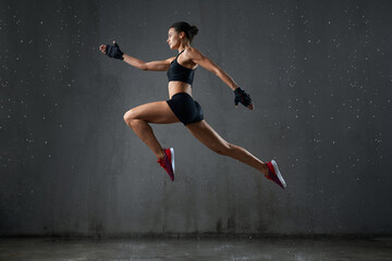 Isolated portrait of athletic woman jumping in hall under rain, loft gray interior. Side view of wet female bodybuilder in sports underwear doing running pose in air during jump. Concept of sport.